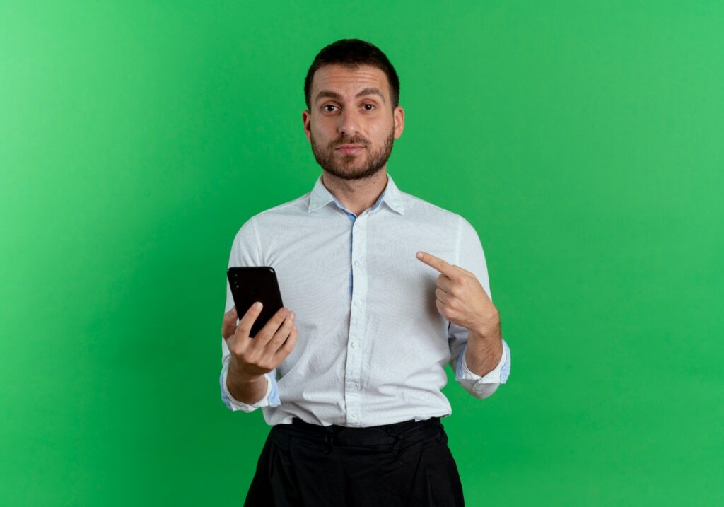 Homem vestindo camisa branca e calças pretas, apontando para um smartphone, contra um fundo verde.