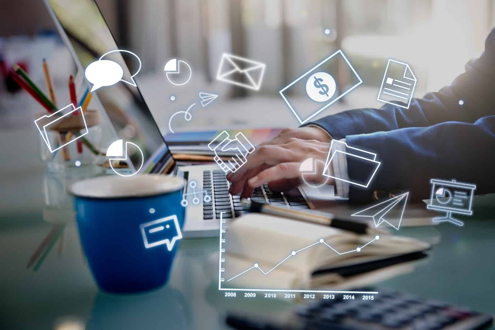 Uma pessoa digitando em um laptop cercada por ícones representativos de comunicação digital e análise de dados, com um copo azul e lápis coloridos ao lado, em um ambiente de escritório